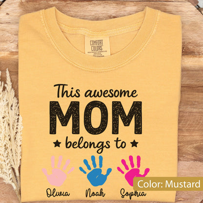 May include: Mustard-colored t-shirt with the text "This awesome MOM belongs to" and three handprints in pink, blue, and pink. The names Olivia, Noah, and Sophia are below the handprints. The shirt is made by Comfort Colors.