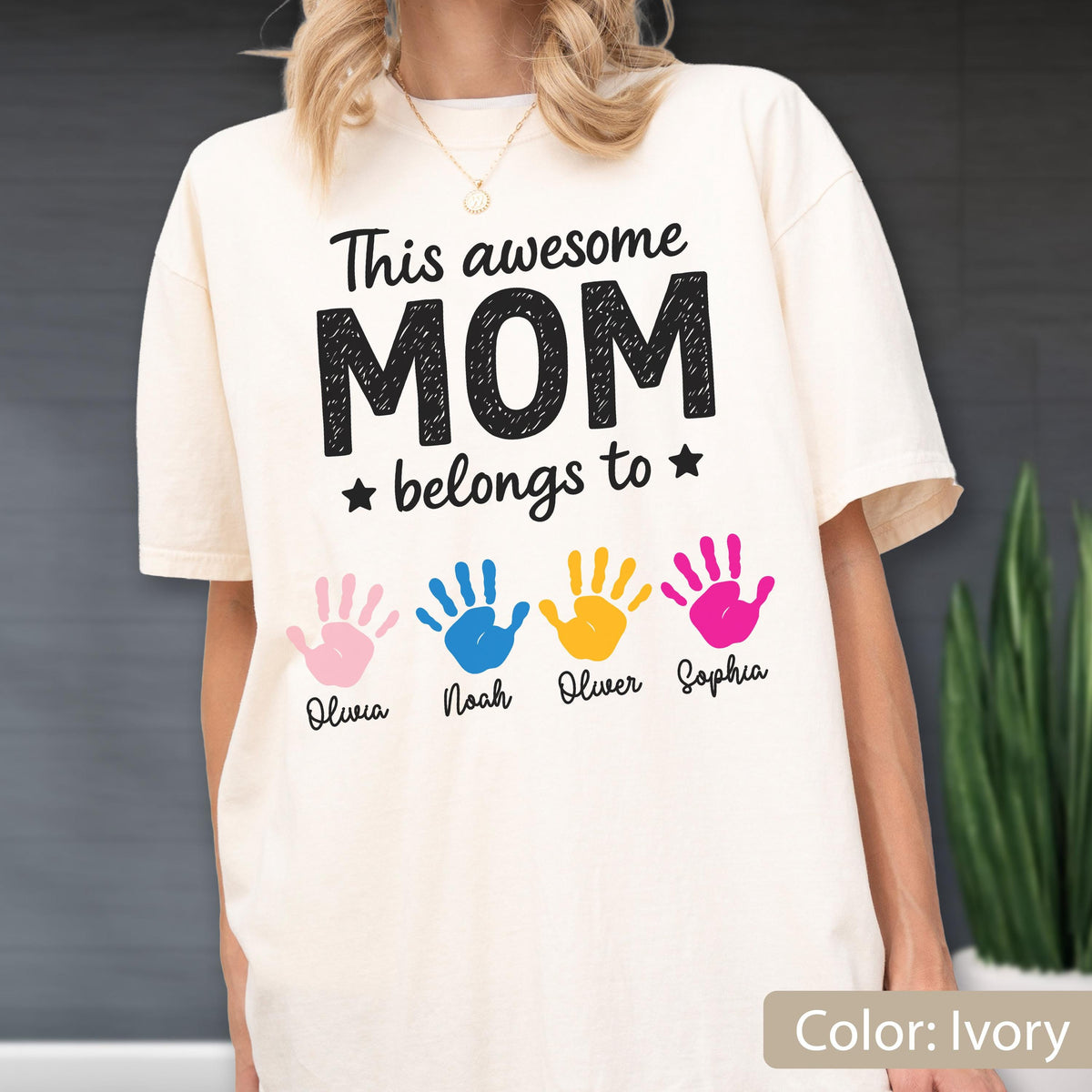 May include: Ivory-colored t-shirt with the text "This awesome MOM belongs to" and four colorful handprints. The names Olivia, Noah, Oliver, and Sophia are printed below the handprints. The shirt is a relaxed fit.