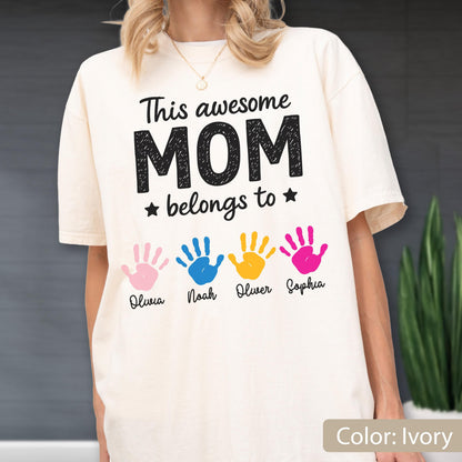 May include: Ivory-colored t-shirt with the text "This awesome MOM belongs to" and four colorful handprints. The names Olivia, Noah, Oliver, and Sophia are printed below the handprints. The shirt is a relaxed fit.
