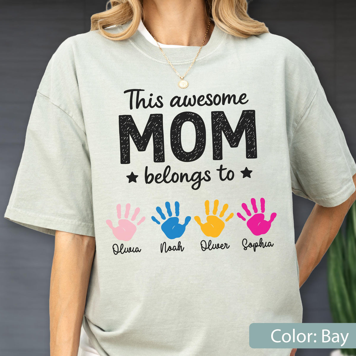 May include: A light green t-shirt with the text "This awesome MOM belongs to" and four colorful handprints. The handprints are pink, blue, yellow, and pink, with names below each.