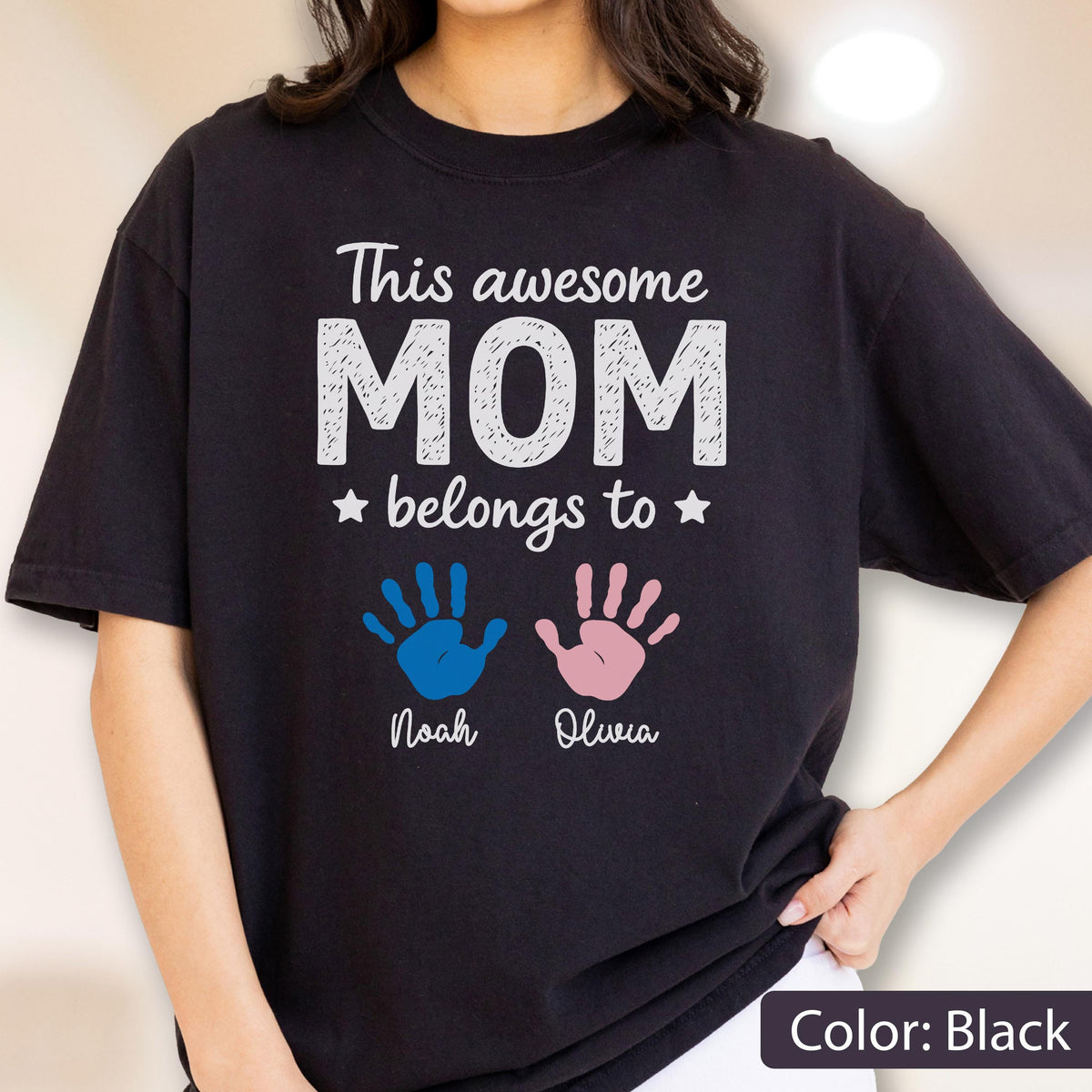 May include: Black t-shirt with the text "This awesome MOM belongs to" above two handprints, one blue and one pink. Below the handprints are the names "Noah" and "Olivia". The shirt is a solid black color.