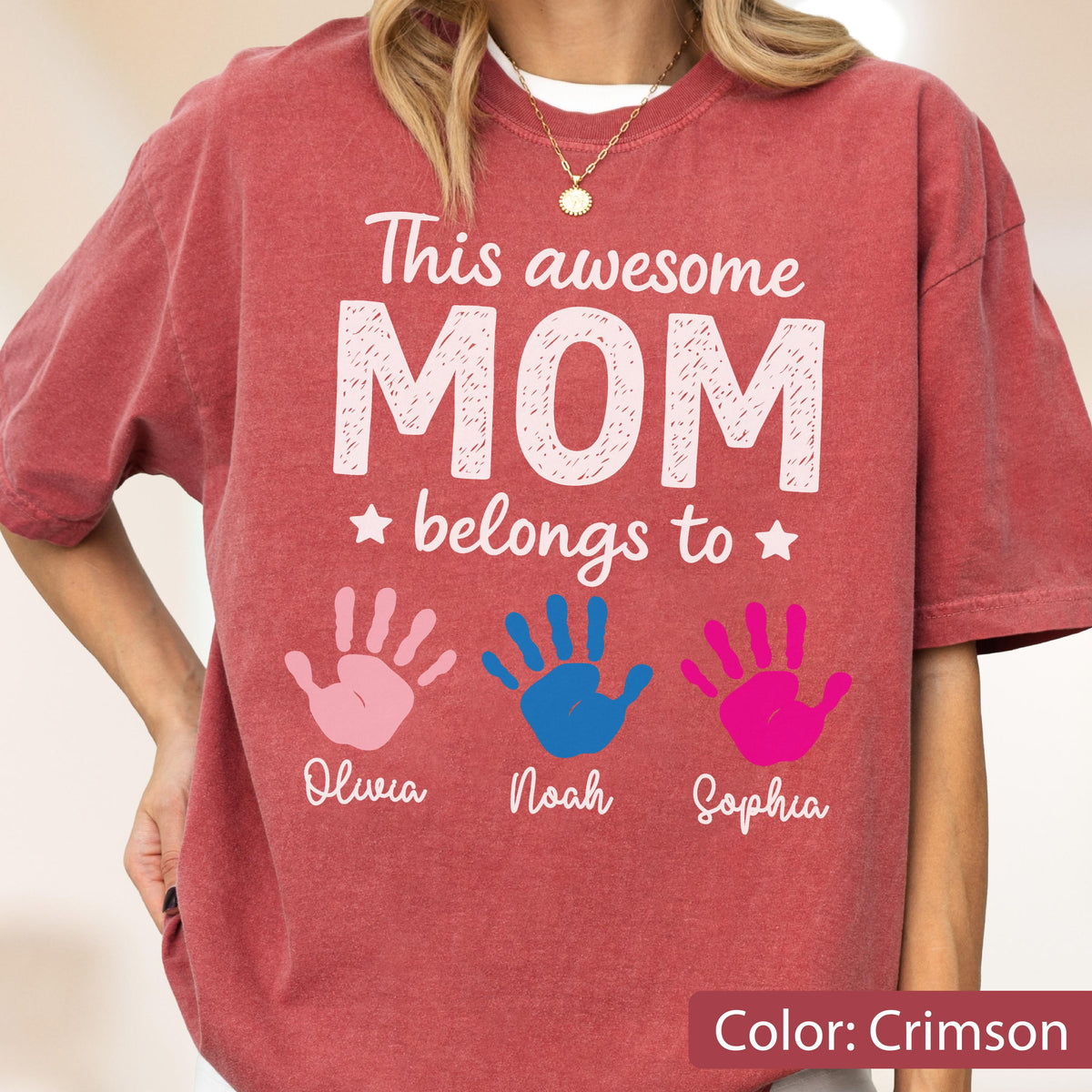 May include: Crimson-colored t-shirt with the text "This awesome MOM belongs to" and three handprints in pink, blue, and magenta. The names Olivia, Noah, and Sophia are printed below the handprints. The shirt is a comfortable, casual style.