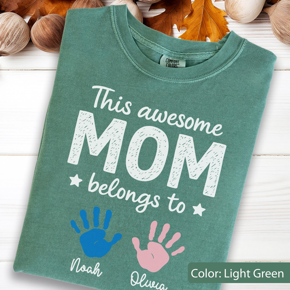 May include: Light green t-shirt with the text "This awesome MOM belongs to" and two handprints, one blue and one pink. The names "Noah" and "Olivia" are printed below the handprints. The shirt is a Comfort Colors brand.