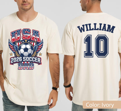 May include: Ivory-colored t-shirt with a soccer-themed graphic. The front features "USA" in red and blue, a soccer ball, wings, and "2026 SOCCER DAD." The back displays "WILLIAM" and the number "10" in navy blue.