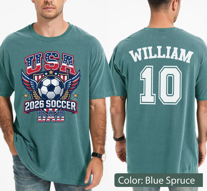 May include: Blue spruce t-shirt with a soccer-themed graphic. The front features "USA" in red, white, and blue, a soccer ball, wings, and "2026 SOCCER DAD." The back displays "WILLIAM" and the number "10" in white.