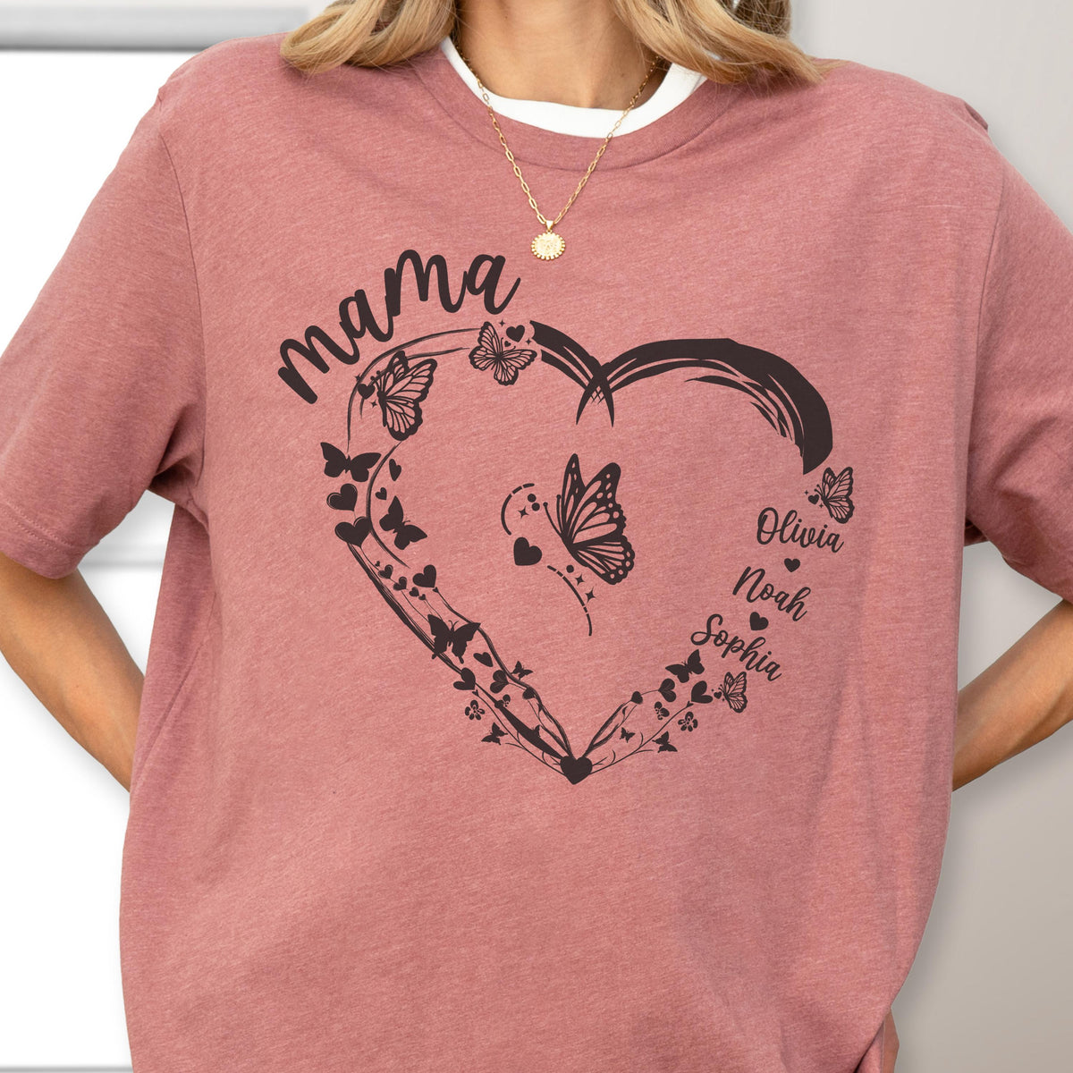 May include: A dusty rose t-shirt featuring a heart-shaped design with butterflies and the word "mama" in a script font. The names Olivia, Noah, and Sophia are printed inside the heart.