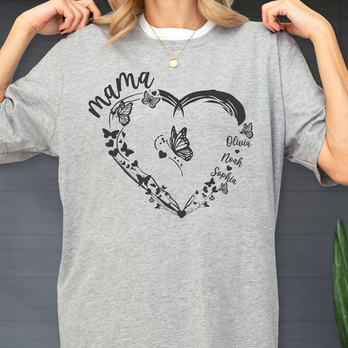 May include: A gray t-shirt with a heart-shaped design featuring butterflies and the names Olivia, Noah, and Sophia. The word "mama" is written above the heart in a cursive font. The shirt has a crew neck.