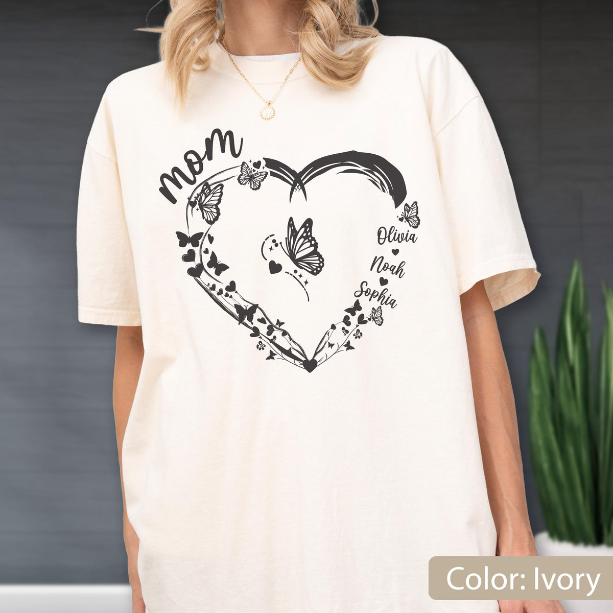 May include: Ivory-colored t-shirt featuring a black heart-shaped design with butterflies and the word "Mom" in script. The names Olivia, Noah, and Sophia are also included. The shirt is a relaxed fit.