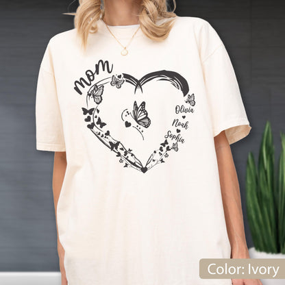May include: Ivory-colored t-shirt featuring a black heart-shaped design with butterflies and the word "Mom" in script. The names Olivia, Noah, and Sophia are also included. The shirt is a relaxed fit.