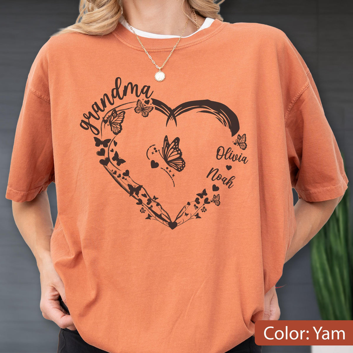 May include: A yam-colored t-shirt featuring a black heart-shaped design with butterflies and the word "grandma" in script. The names "Olivia" and "Noah" are also included. The shirt is a casual style.