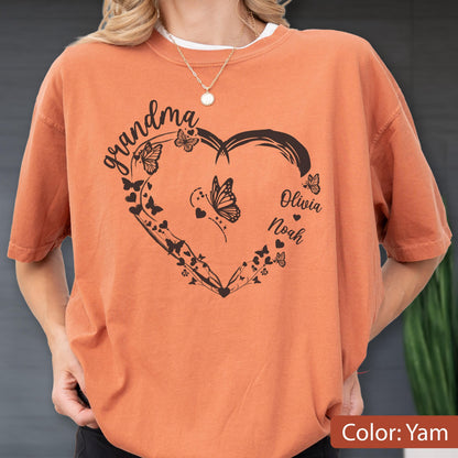 May include: A yam-colored t-shirt featuring a black heart-shaped design with butterflies and the word "grandma" in script. The names "Olivia" and "Noah" are also included. The shirt is a casual style.