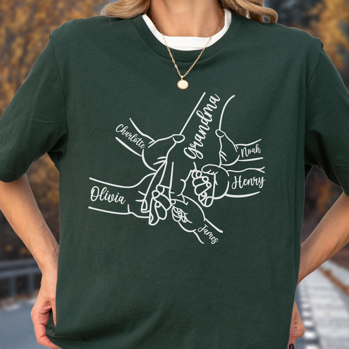 May include: Dark green t-shirt with a white graphic of hands holding each other, with the word "Grandma" and names "Charlotte", "Olivia", "Noah", "Henry", and "James". The shirt has a white crew neck.