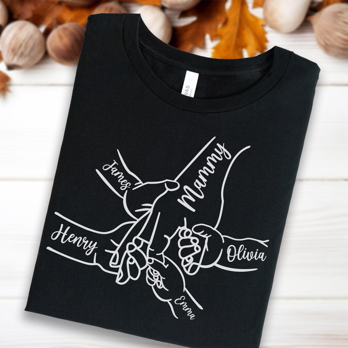 May include: Black t-shirt with a white graphic of hands holding each other, with the names "James", "Mammy", "Henry", "Olivia", and "Emma" printed on the shirt. The shirt is folded and displayed on a light wood surface.