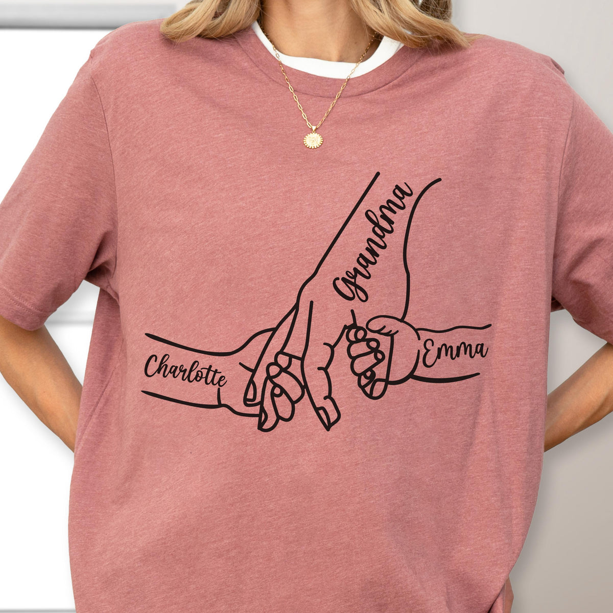 May include: Dusty rose t-shirt featuring a black graphic of hands holding each other. The word "Grandma" is written above the largest hand, with "Charlotte" and "Emma" written on the other hands.