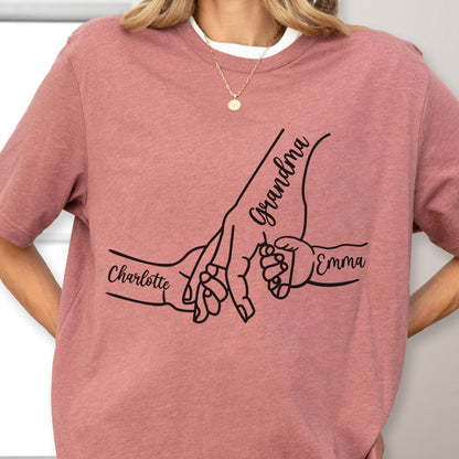 May include: Dusty rose t-shirt featuring a black graphic of hands holding each other. The word "Grandma" is written above the largest hand, with "Charlotte" and "Emma" written on the other hands.