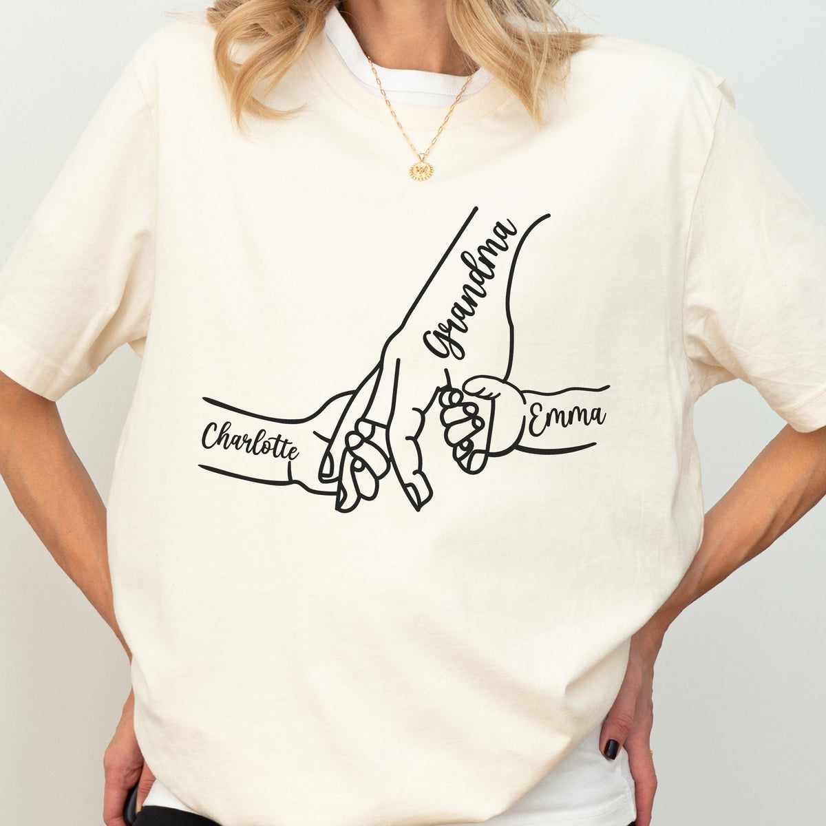 May include: Cream-colored t-shirt featuring a black line drawing of hands holding each other. The word "Grandma" is written above the larger hand, with "Charlotte" and "Emma" written on either side.
