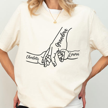 May include: Cream-colored t-shirt featuring a black line drawing of hands holding each other. The word "Grandma" is written above the larger hand, with "Charlotte" and "Emma" written on either side.