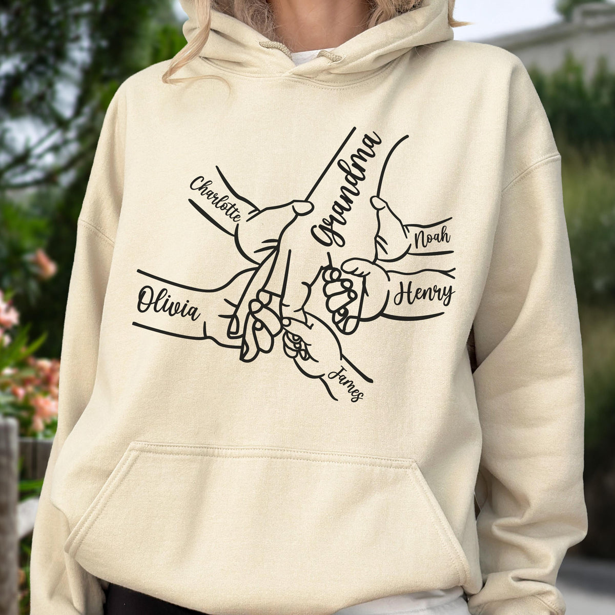 May include: A beige hoodie featuring a black graphic of hands intertwined, with the word "Grandma" and the names "Charlotte", "Olivia", "James", "Noah", and "Henry" printed on it. The hoodie has a front pocket and a hood.