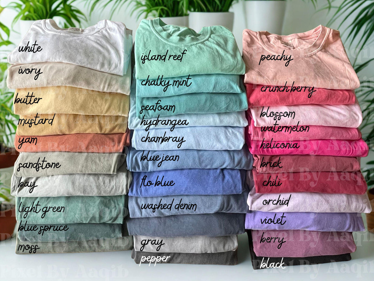 May include: Stacks of folded t-shirts in various colors, including white, island reef, and peachy. Each shirt is labeled with its color name in handwritten script. The shirts are neatly arranged, showcasing a range of hues from light to dark.
