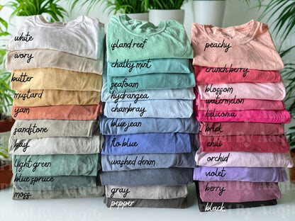 May include: Stacks of folded t-shirts in various colors, including white, island reef, and peachy. Each shirt is labeled with its color name in handwritten script. The shirts are neatly arranged, showcasing a range of hues from light to dark.