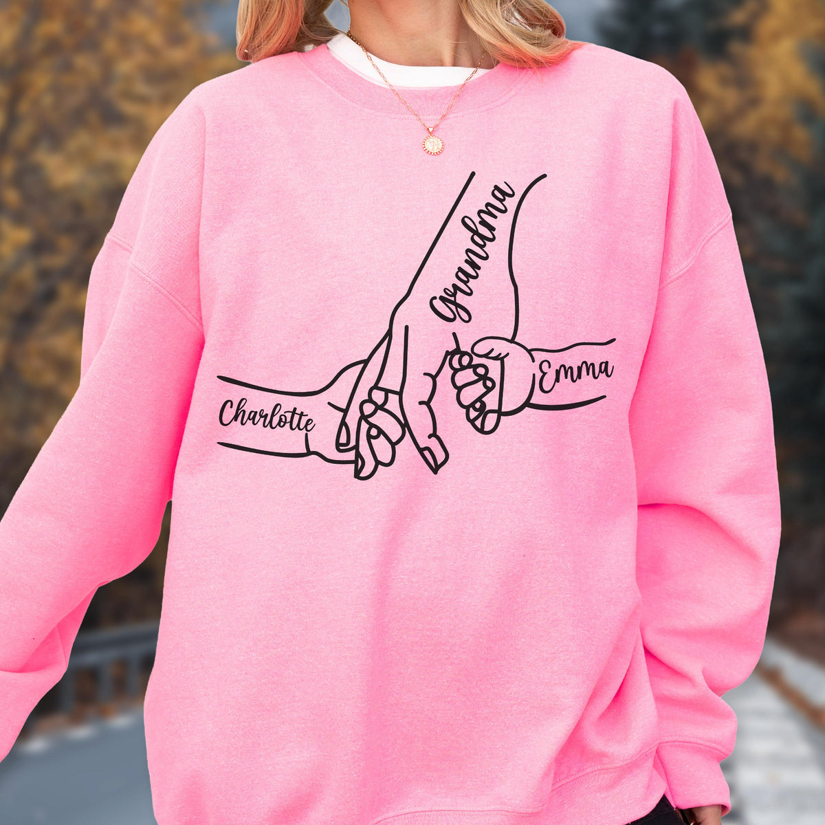 May include: Pink sweatshirt featuring a black line drawing of a grandmother's hand holding a child's hand, with the names "Charlotte" and "Emma" on the sleeves. The word "Grandma" is written on the grandmother's hand.