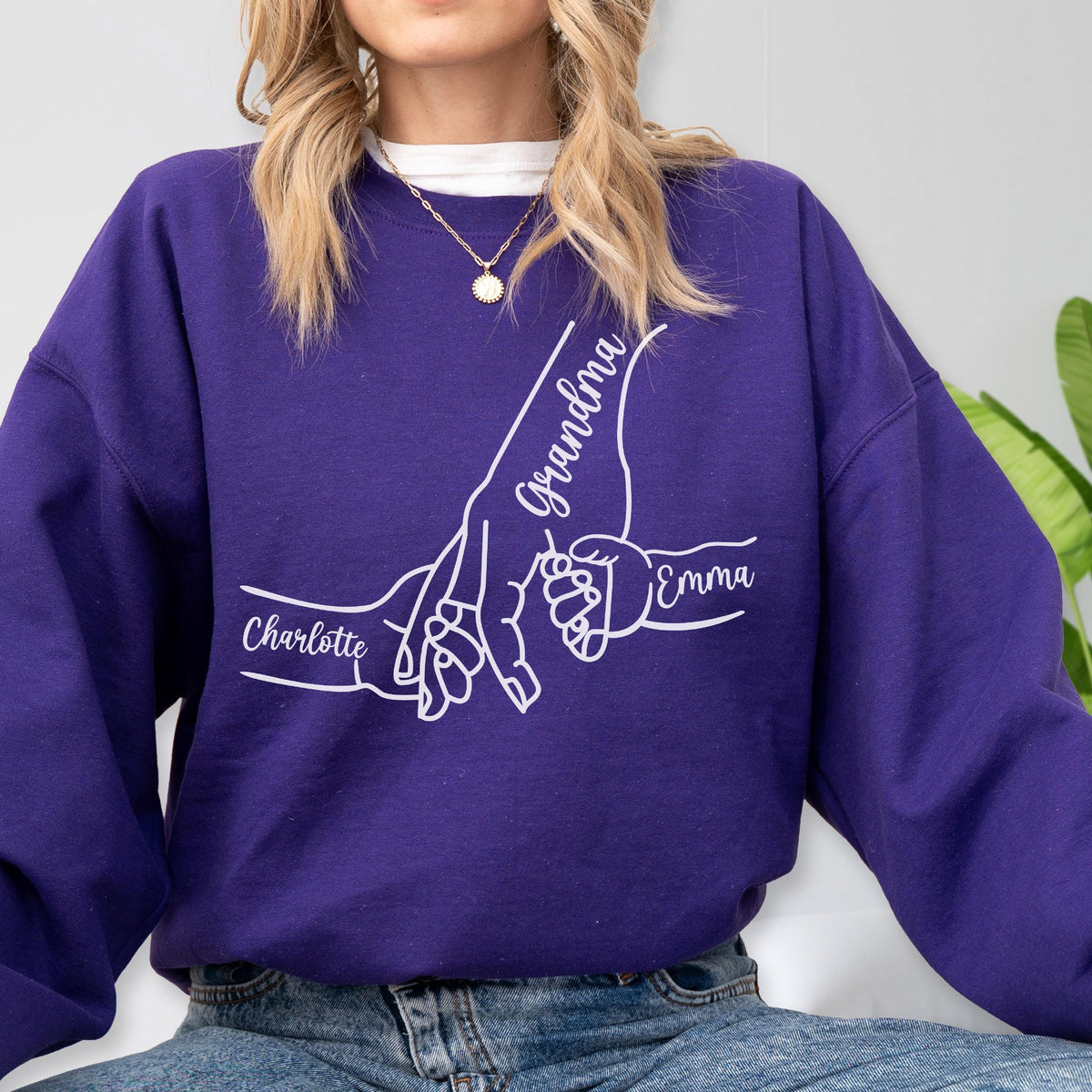May include: A purple sweatshirt with a white graphic of hands holding each other. The word "Grandma" is written on one hand, and the names "Charlotte" and "Emma" are on the other hands. The sweatshirt is made of a soft material.