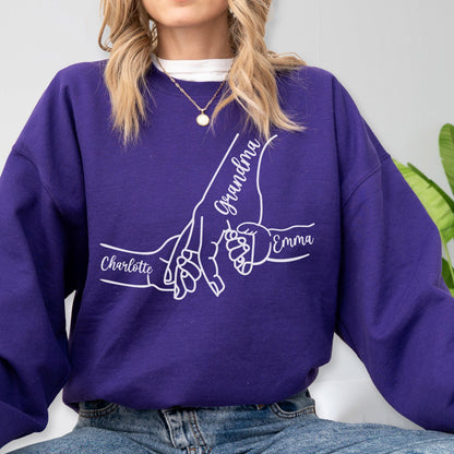 May include: A purple sweatshirt with a white graphic of hands holding each other. The word "Grandma" is written on one hand, and the names "Charlotte" and "Emma" are on the other hands. The sweatshirt is made of a soft material.