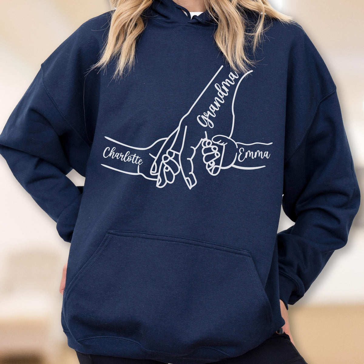 May include: Navy blue hoodie with a white graphic of hands holding each other. The word "Grandma" is written in cursive, with the names "Charlotte" and "Emma" also displayed. The hoodie has a front pocket and a hood.
