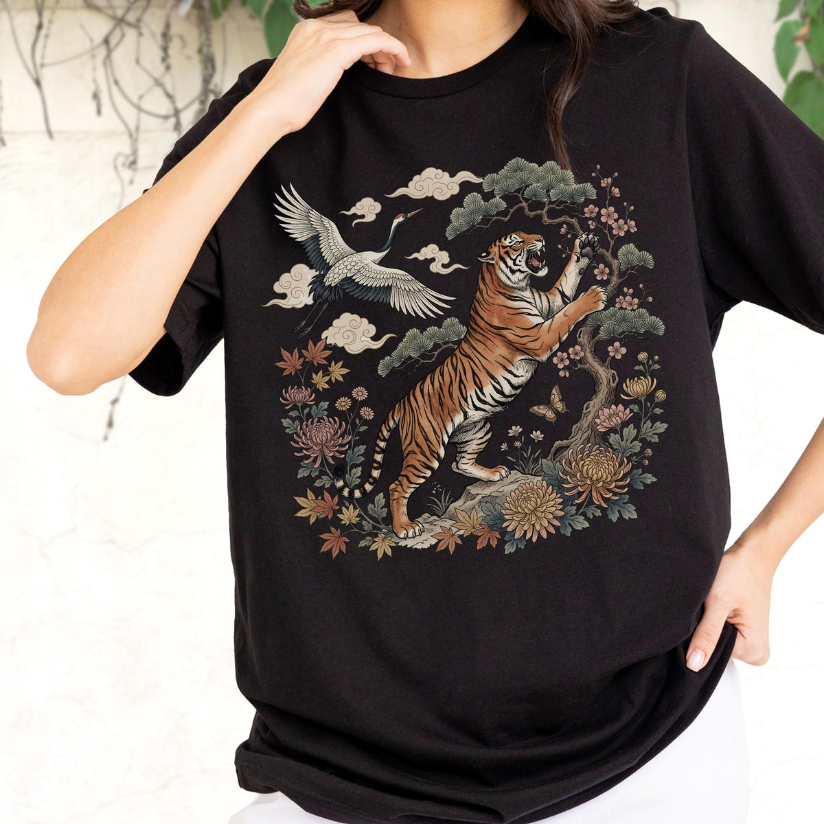 May include: Black t-shirt featuring a detailed illustration of a tiger, crane, and floral elements. The tiger is depicted in a dynamic pose, set against a backdrop of stylized clouds and foliage. The design incorporates shades of orange, brown, and green.
