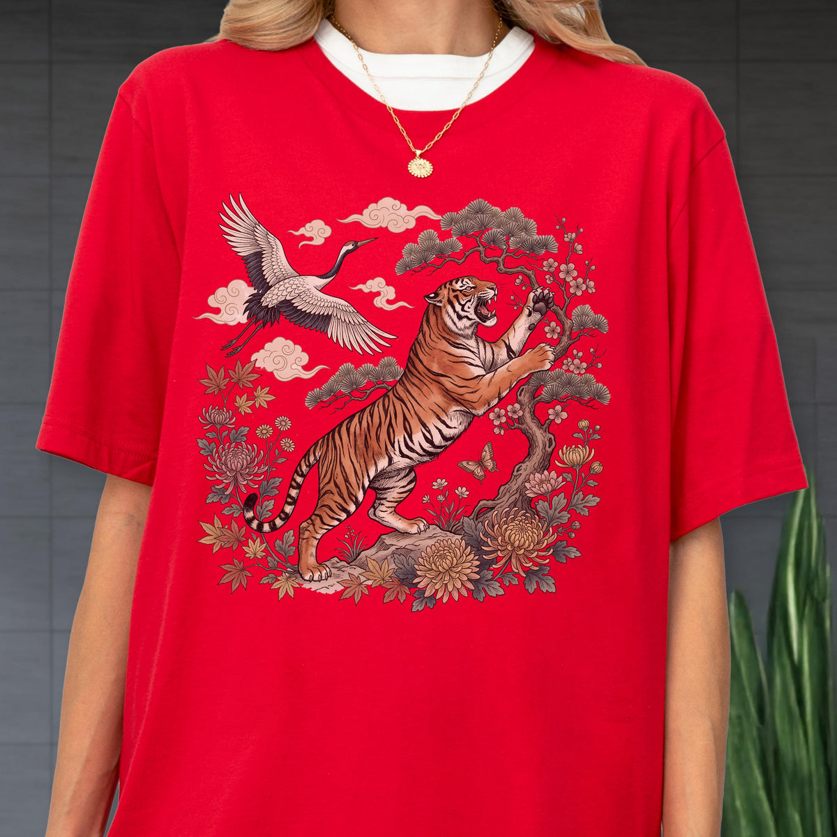 May include: Red t-shirt featuring a detailed illustration of a tiger, crane, and floral elements. The tiger is depicted with orange and black stripes, reaching towards a tree. The design includes clouds and flowers, creating a vibrant and artistic composition.