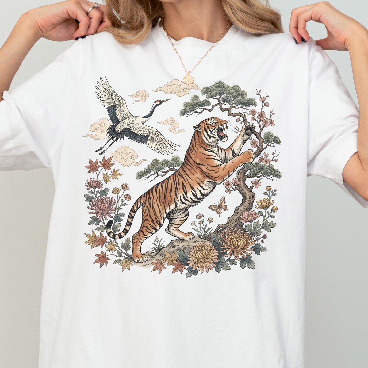 May include: White t-shirt featuring a detailed illustration of a tiger, crane, and floral elements. The tiger is depicted with orange and black stripes, reaching towards a tree. A crane flies above, with clouds in the background.