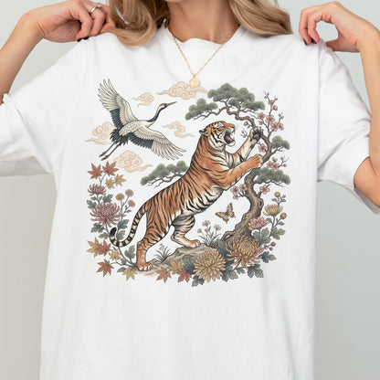 May include: White t-shirt featuring a detailed illustration of a tiger, crane, and floral elements. The tiger is depicted with orange and black stripes, reaching towards a tree. A crane flies above, with clouds in the background.