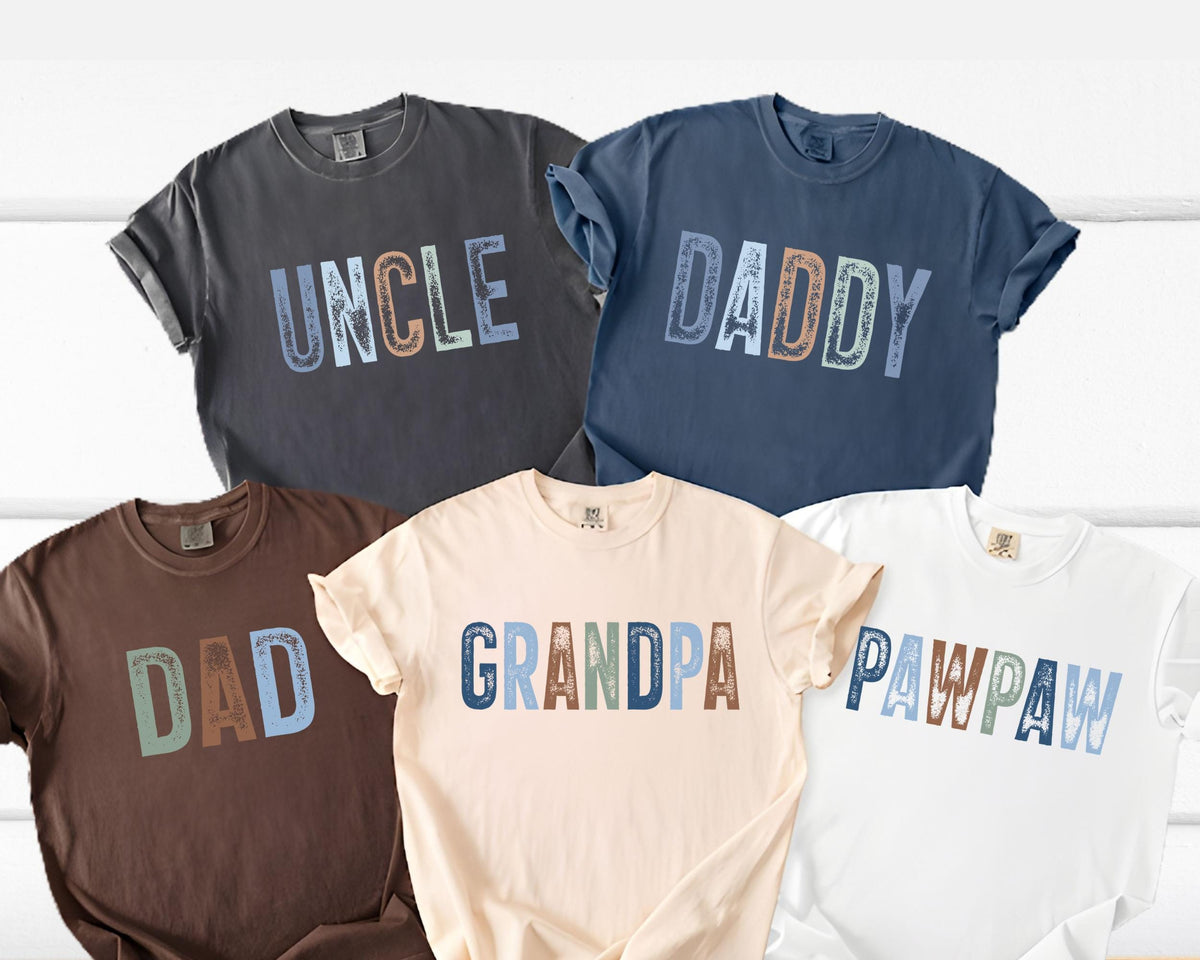 May include: Five t-shirts in various colors, each with a different word printed across the front: "UNCLE", "DADDY", "DAD", "GRANDPA", and "PAWPAW". The text is in a multi-colored, distressed font. The shirts are rolled up at the sleeves.