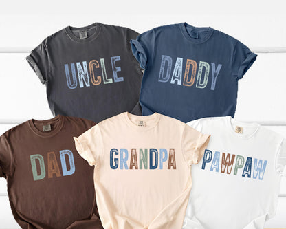 May include: Five t-shirts in various colors, each with a different word printed across the front: "UNCLE", "DADDY", "DAD", "GRANDPA", and "PAWPAW". The text is in a multi-colored, distressed font. The shirts are rolled up at the sleeves.
