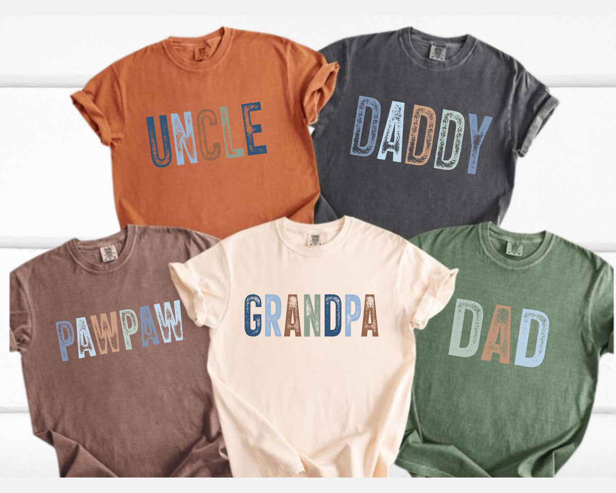 May include: Five short-sleeved t-shirts in various colors, each with a different word printed on the front. The words are "UNCLE", "DADDY", "PAWPAW", "GRANDPA", and "DAD", in a multi-colored, block letter design.
