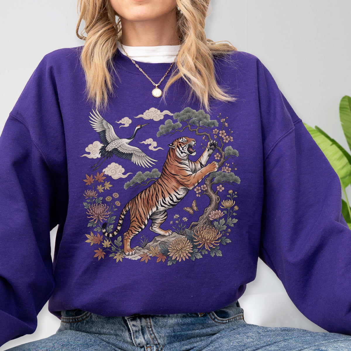May include: A purple sweatshirt featuring a detailed illustration of a tiger, crane, and floral elements. The tiger is depicted in shades of orange and black, set against a backdrop of stylized clouds and flowers. The design is centered on the front of the sweatshirt.