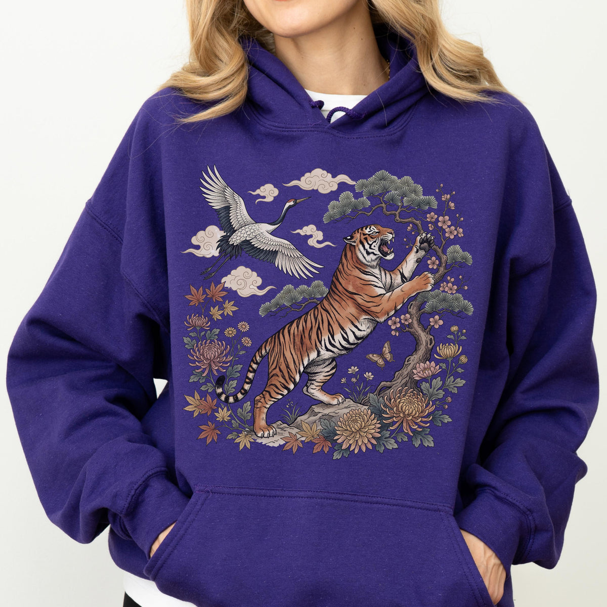 May include: A purple hoodie featuring a detailed illustration of a tiger, crane, and floral elements. The tiger is depicted with orange and black stripes, reaching towards a flowering tree. A crane flies above, with clouds and flowers completing the design.