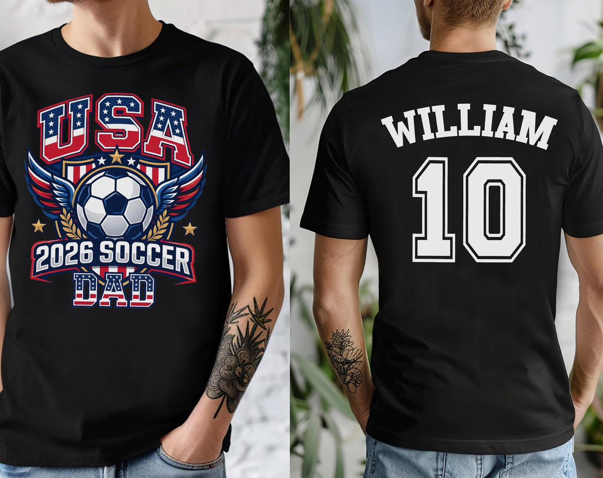 May include: Black t-shirt with a soccer-themed graphic. The front features "USA" in red and blue, a soccer ball, wings, and "2026 SOCCER DAD." The back displays "WILLIAM" and the number "10" in white.