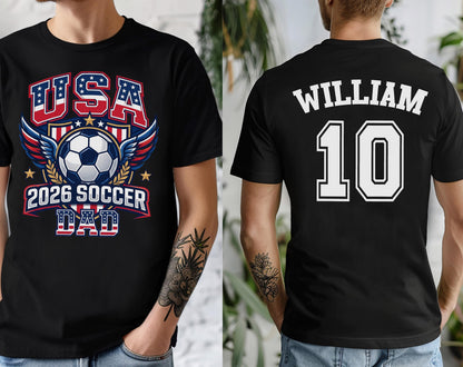 May include: Black t-shirt with a soccer-themed graphic. The front features "USA" in red and blue, a soccer ball, wings, and "2026 SOCCER DAD." The back displays "WILLIAM" and the number "10" in white.