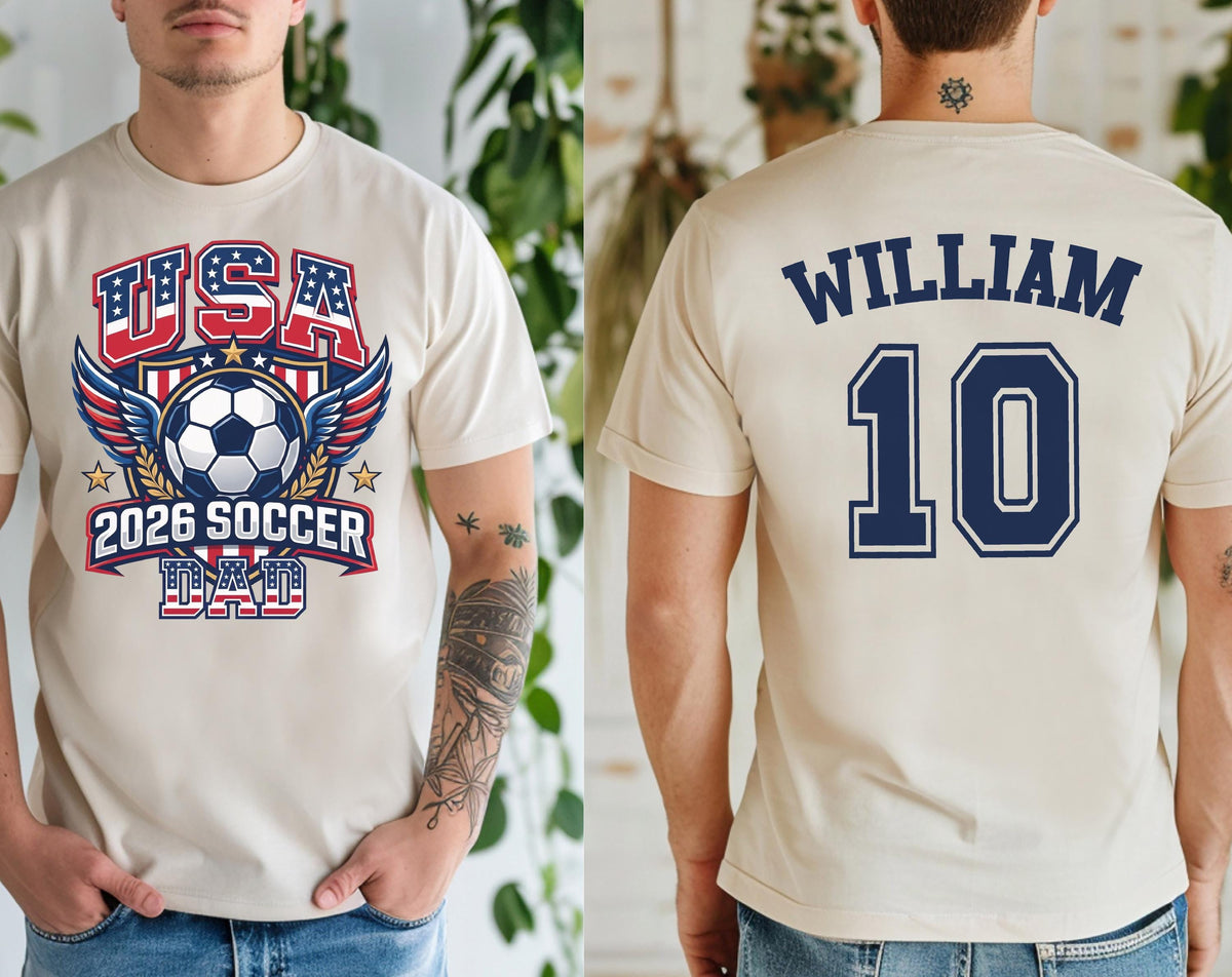 May include: Beige t-shirt with a soccer-themed graphic. The front features "USA" in red and blue, a soccer ball, and "2026 SOCCER DAD." The back displays "WILLIAM" and the number "10" in navy blue.