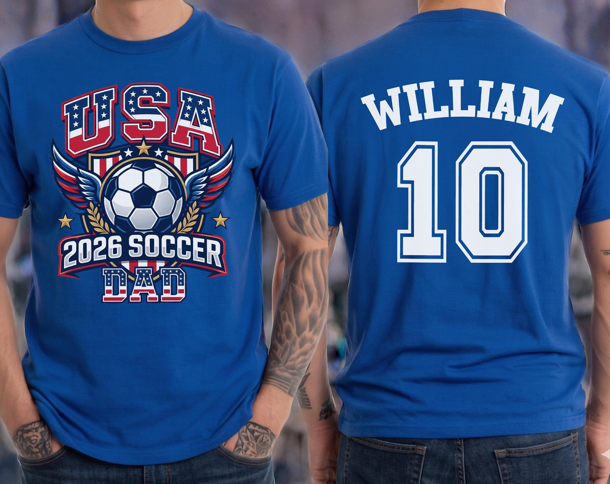 May include: Royal blue t-shirt with a soccer-themed graphic on the front, featuring the text "USA 2026 SOCCER DAD" and a soccer ball design. The back of the shirt displays the name "WILLIAM" and the number "10" in white.