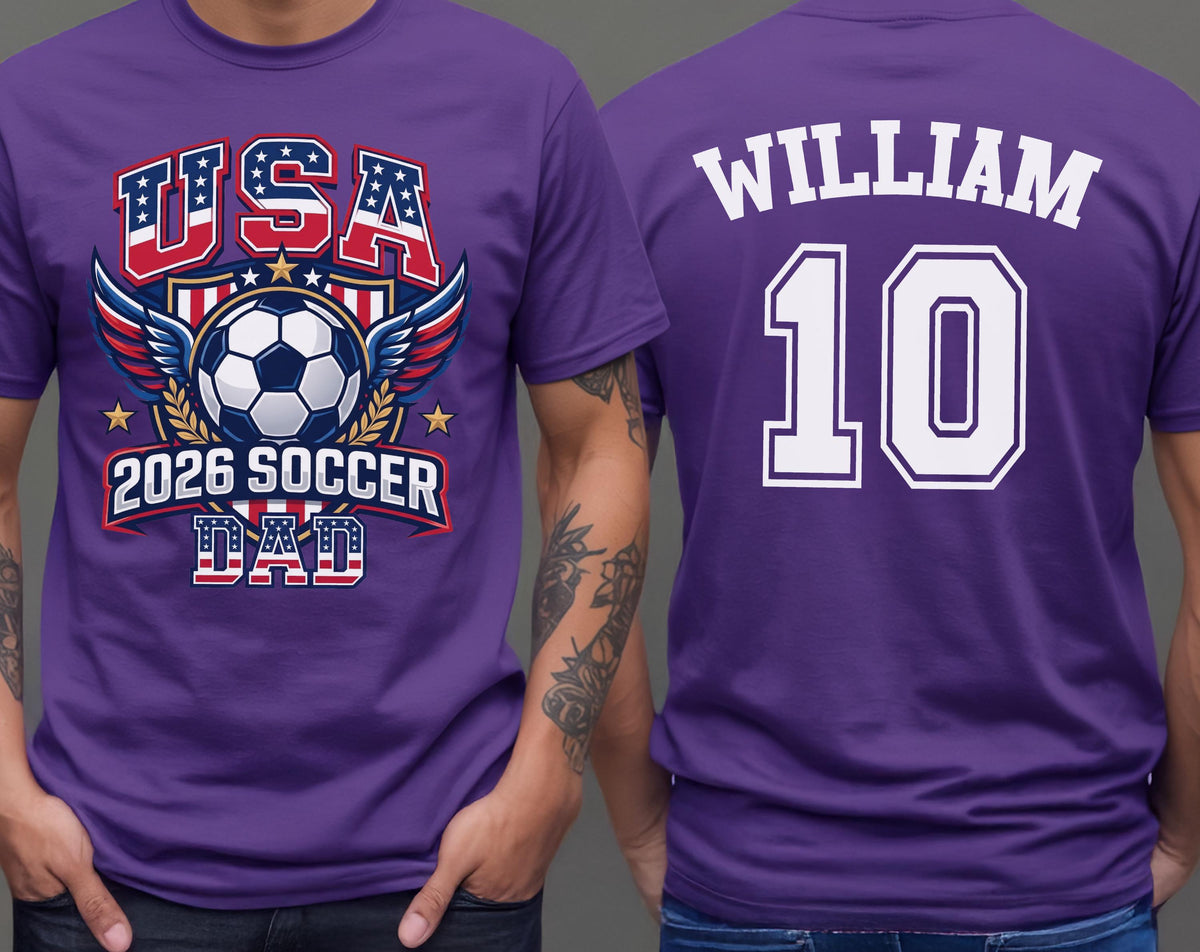May include: Purple t-shirt with a soccer-themed graphic. The front features "USA" in red and blue, a soccer ball, and "2026 SOCCER DAD." The back displays "WILLIAM" and the number "10" in white.