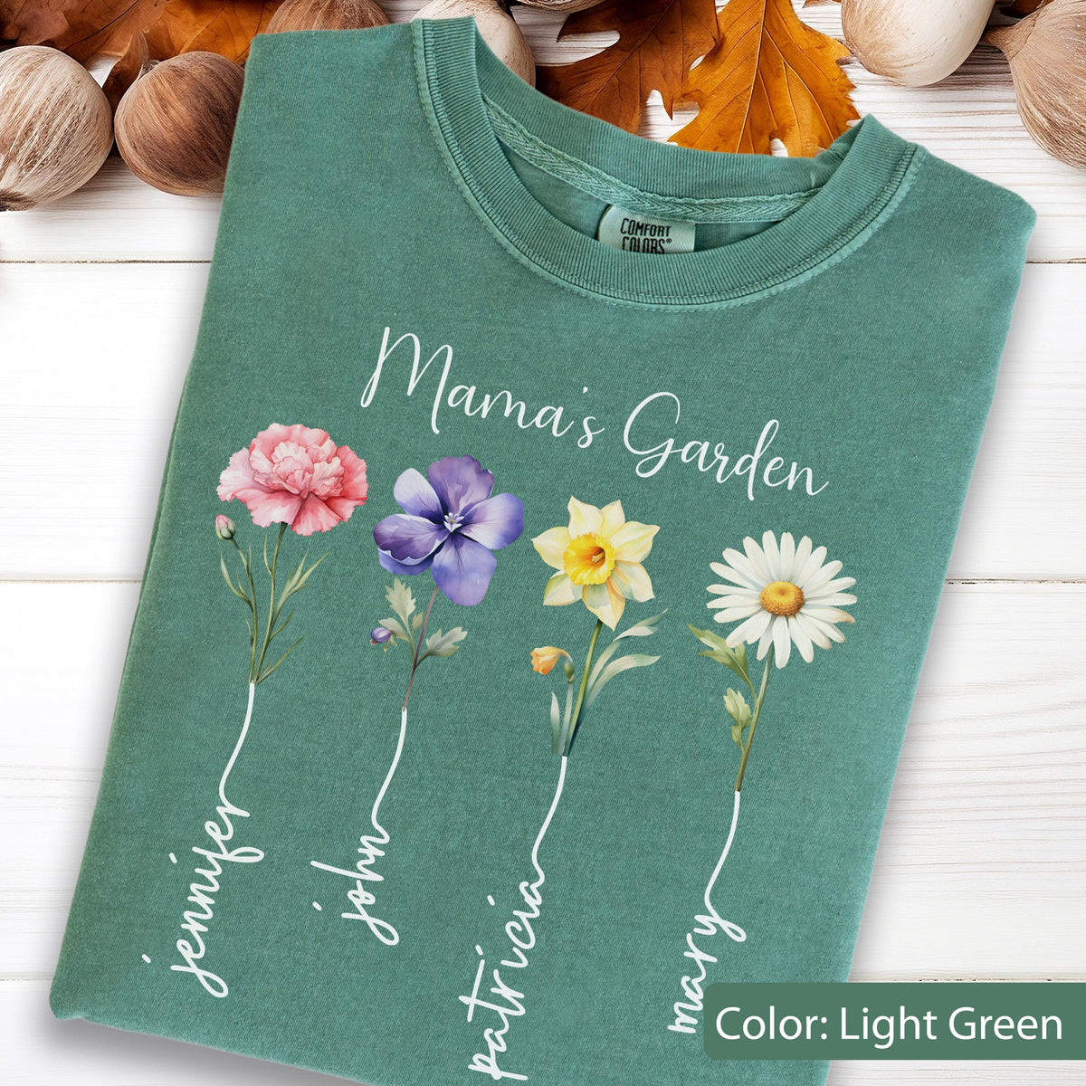 May include: Light green t-shirt with the text "Mama's Garden" and floral illustrations. The shirt features a pink carnation, purple pansy, yellow daffodil, and white daisy. Names are written below each flower: Jennifer, John, Patricia, and Mary.