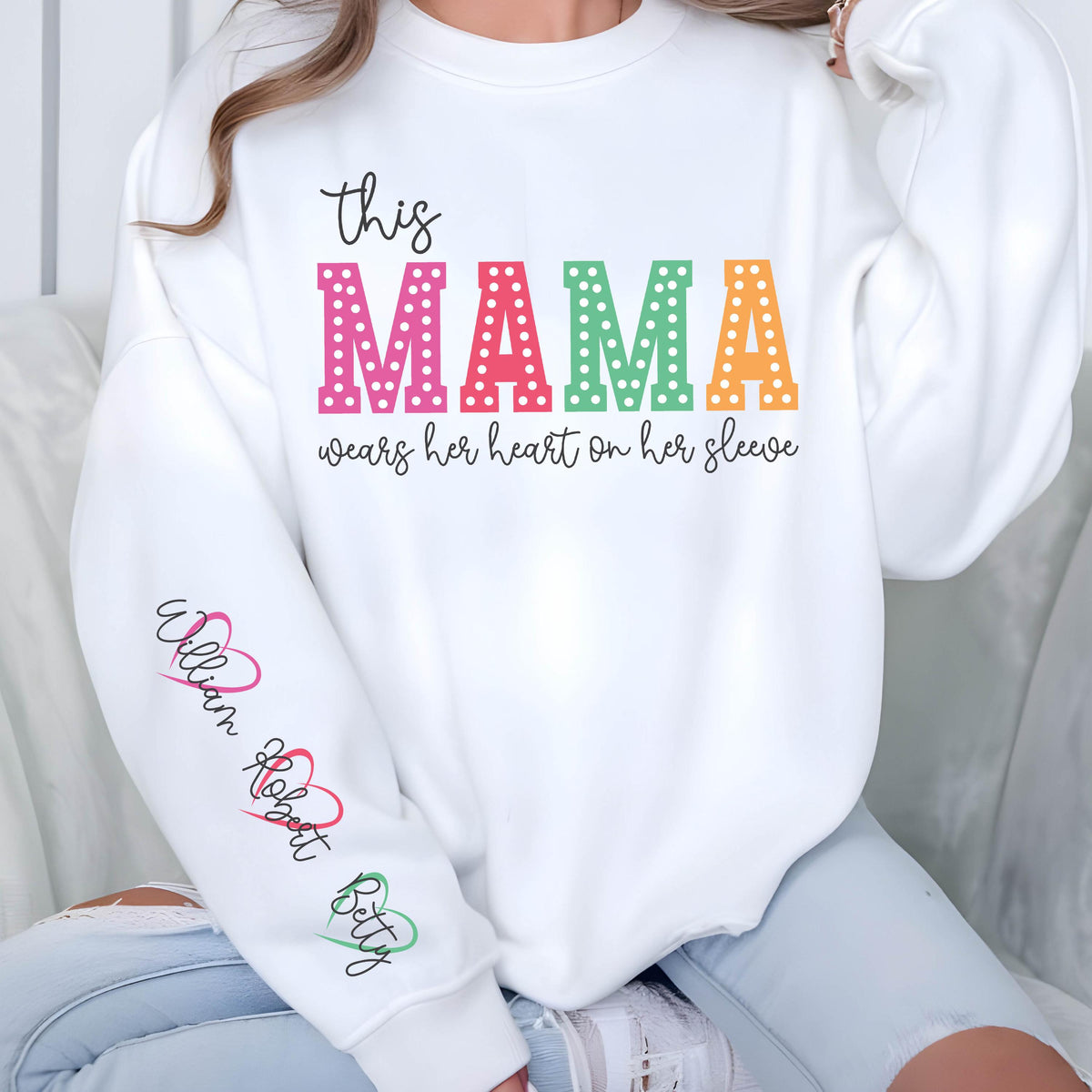 May include: White sweatshirt with the text "this MAMA wears her heart on her sleeve" in rainbow colors with white polka dots. The sleeves have the names "William", "Robert", and "Betty" written in cursive with hearts.
