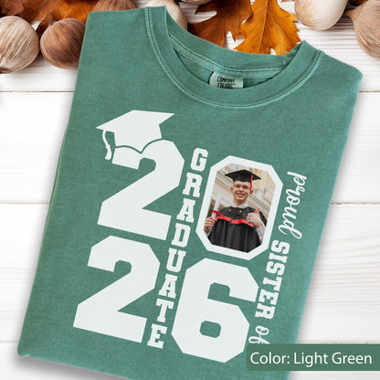 May include: Light green t-shirt with the text "2026 Graduate" and a photo of a graduate. The words "proud sister of" are printed vertically on the right side. The shirt has a crew neck and short sleeves.