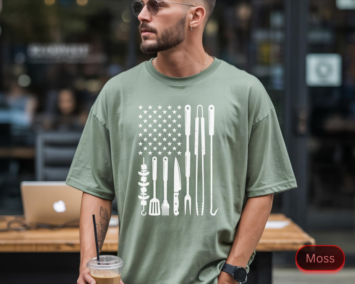 May include: A moss green t-shirt featuring a white graphic design of the American flag with grilling tools. The design includes stars, skewers, a spatula, knife, fork, and tongs. The shirt is a relaxed fit.