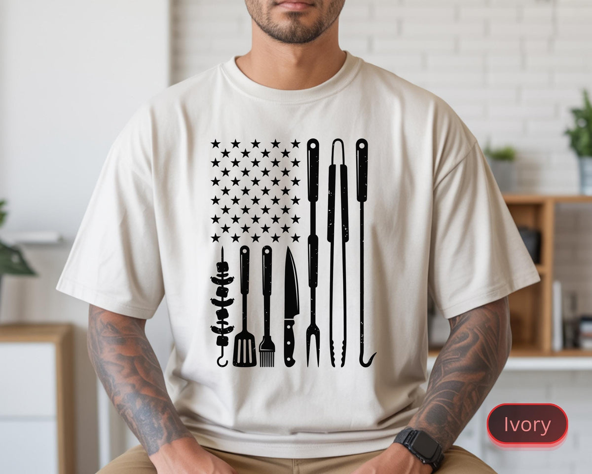 May include: Ivory-colored t-shirt featuring a black graphic design of the American flag with grilling tools replacing the stripes. The design includes stars, skewers, a spatula, a fork, a knife, and grilling hooks. The shirt is a casual, short-sleeved style.