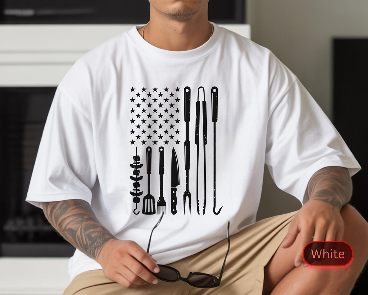 May include: White t-shirt featuring a black graphic design of an American flag made of grilling tools. The design includes stars and various barbecue utensils, such as skewers, spatulas, and knives. The shirt is a casual, short-sleeved style.