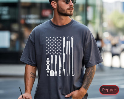 May include: Dark gray t-shirt featuring a white graphic design of the American flag, with the stars replaced by a pattern and the stripes by grilling tools. The word "Pepper" is in a red oval on the bottom right.