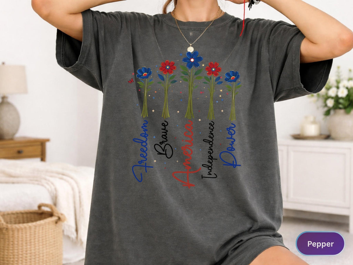 May include: Dark gray t-shirt featuring a floral design with the words "Freedom, Brave, America, Independence, Power" in red and blue script. The flowers have red and blue petals and green stems.